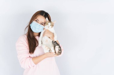 Koronavirüsü korumak için tıbbi koruma maskesi takan Asyalı güzel mutlu kadın başbakan 2.5 numaralı filtre tozu, kedi kürküne alerjisi var ve beyaz arka planda bir kedi taşıyor.