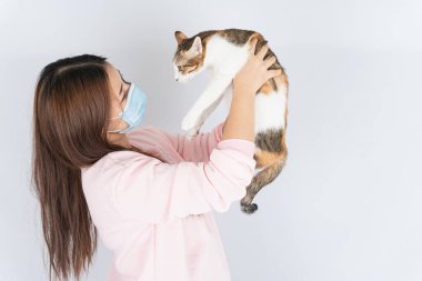 Koronavirüsü korumak için tıbbi koruma maskesi takan Asyalı güzel mutlu kadın başbakan 2.5 numaralı filtre tozu, kedi kürküne alerjisi var ve beyaz arka planda bir kedi taşıyor.