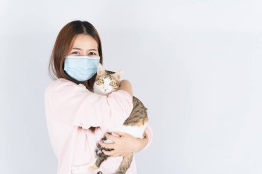 Koronavirüsü korumak için tıbbi koruma maskesi takan Asyalı güzel mutlu kadın başbakan 2.5 numaralı filtre tozu, kedi kürküne alerjisi var ve beyaz arka planda bir kedi taşıyor.