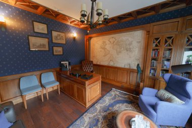 Büyük meşe masalı klasik bir çalışma. Misafirler için deri kanepe. Duvarda kocaman bir dünya haritası var. İç kısımda viski masası ve papağan heykeli de var. Avize kapalı.