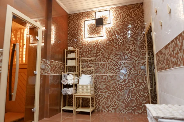 The interior of the dressing room, the entrance to the Russian bathhouse. Part of the wall is made of red and white mosaic squares. Part of the wall is lined with ceramic tiles. On the wall is a beautifully styled lamp in the form of two squares. Lef