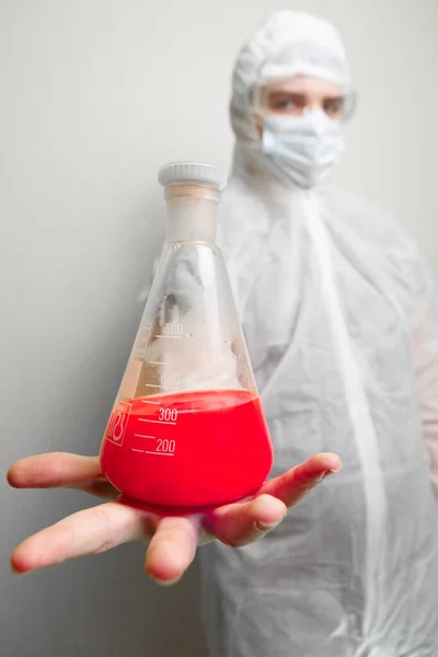 Coronovirus pandemic in the world 2020. Science against coronovirus. In the frame, a laboratory assistant in a white suit, hat and mask. Holds a flask in his hand with a red liquid symbolizing coronovirus. Flask in the foreground.