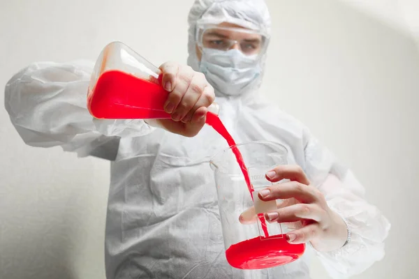 Coronovirus pandemic in the world 2020. Science against coronovirus. In the frame, a laboratory assistant in a white suit, hat and mask. Holds a flask in his hand with a red liquid symbolizing coronovirus. Flask in the foreground.