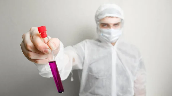 Science against coronovirus. In the frame, a laboratory assistant in a white suit, hat and mask. Holds a purple liquid in a test tube in the foreground. On a white background.