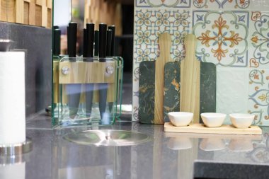 Mutfak gereçleri: bıçaklar, kesim tahtaları, bir rulo peçete, sos kaseleri. Taş bir tezgahın üzerinde dur. Duvar seramik fayanslarla süslenmiş..