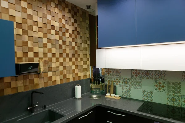 Modern kitchen in black and blue, corner with a wooden wall. Corner of the kitchen with sink, stove, wall cabinet. The backlight is on. There are kitchen utensils on the countertop.