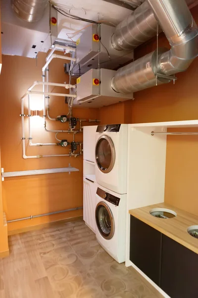 Utility room with two washing machines. There are large pipes on the ceiling. There are many water pipes on the walls. The lamp is on.