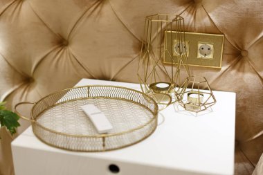 Altın renkli dekoratif elementler beyaz bir komodinin üzerinde yatıyor. Uzaktan kumanda için dur. Askılar. Duvar yumuşak kumaşla süslenmiş. Çerçevedeki oyuklar.