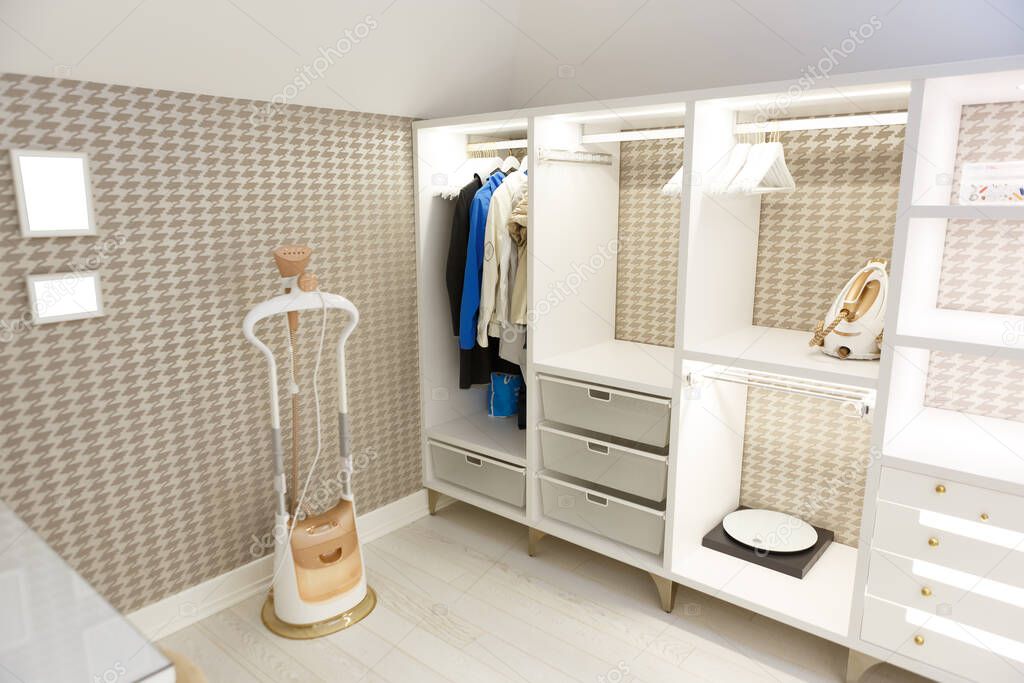 Beige wardrobe for clothes in the dressing room. Open shelves, things hang on a hanger. On the shelves, scales, iron, hangers. On the floor is a garment steamer. Picture Frames.