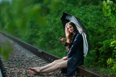 Ormandaki tren raylarında siyah şemsiyeli bir kız. Yağmur yağıyor. Kız rayların üzerinde oturuyor, bacakları öne doğru uzanıyor. Saçı gevşek, kameraya bak. Kız parlak ışıkla aydınlatılıyor. Çevrede parlak yeşil orman.