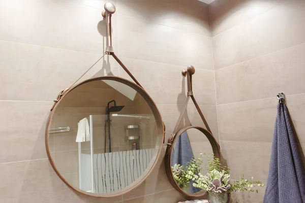 Two round mirrors in the bathroom are trimmed with leather. The reflection of the shower and towels. On the bedside table is a decorative plant. Bathroom in beige colors.