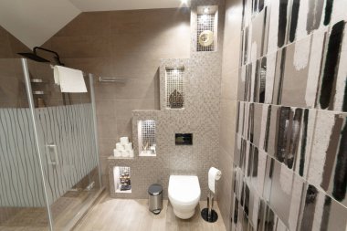 Bej renkli giyinme odası. Çerçevenin içinde duş, tuvalet, tuvalet kağıdı ve havlu var. Duvar ışıklarla süslenmiş, raflarda bir sürü ıvır zıvır var. Dekor elementleri, ahşap figürler, tuvalet fırçası.