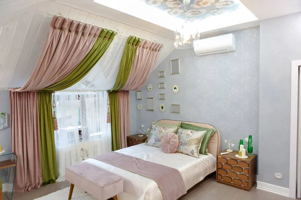 Bedroom in bright colors. On the window are green-pink curtains. Air conditioning on the wall, photo frames on the wall. Dressing table with mirrors. Green armchair. Bed with ataman. The chandelier is shining.