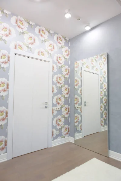 Corner of a room with a door and a large mirror on the wall. Wallpaper of flowers, gray color snten. The door is reflected in the mirror. Interior design of a bedroom. Vertical orientation.