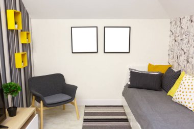 Gri kanepeli yatak odası. Gri koltuk, koltukta parlak yastıklar. Yer paspası. Duvarda iki tane fotoğraf çerçevesi var. Fotoğraflarını koyabilirsin. Sarı dekoratif raflar, çizgili bir duvarda çiçekler..