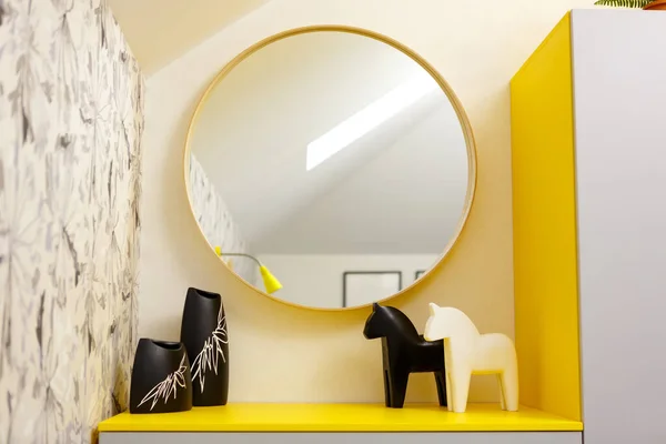 Interior design of a bedroom in yellow tones. Yellow bedside table with figures of horses and vases. Large round mirror on the wall. Lamp and armchair in the foreground in blur. Bright yellow color.