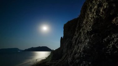 Gece gökyüzünde deniz üzerinde uçan bulutlar ve dolunay arka planında kayalıklar. Zaman aşımı.