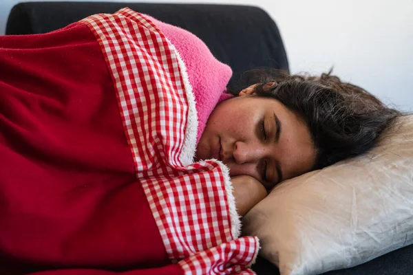 Detail of the face of a woman with long hair in a pink sweater stretched out on a sofa sleeping a nap with a red wool blanket over her during the quarantine of covid-19