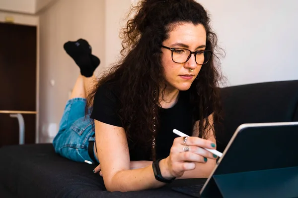 Young brunette girl working from home with her tablet with a digital pencil while lying on a black couch and wearing eyeglasses and blue jeans