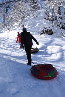 Kızaklı bir adam kızaklı küçük bir kızın peşinden yokuş yukarı tırmanır. İnsanlar karlı bir tepeye tırmanıp binmek için kızak çekerler..