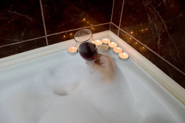 在浴室里 在褐色瓷砖中间 蜡烛在一个女人的手里燃烧着一杯葡萄酒 到处都是浴盆的泡沫 女孩在浴室里 喝一杯葡萄酒和燃烧的蜡烛 图库图片
