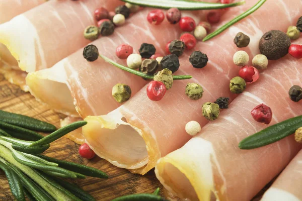 Närbild av tunna skivor prosciutto med rosmarin och paprika på trä skärbräda — Stockfoto