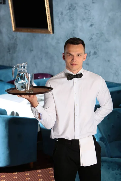 A young, handsome waiter is holding a tray in a carafe of water. The concept of the restaurant business. The staff in the field of restaurant business.