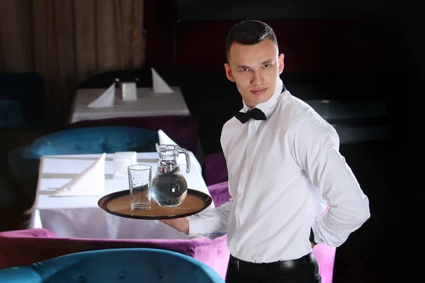 Un camarero joven y guapo sostiene una bandeja en una jarra de agua. El concepto del negocio de los restaurantes. El personal en el campo de los negocios de restaurantes. Fotos en el interior . — Foto de Stock