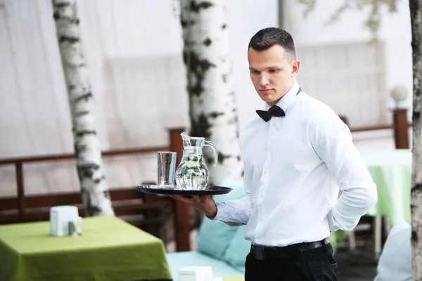 Un camarero joven y guapo sostiene una bandeja en una jarra de agua. El concepto del negocio de los restaurantes. El personal en el campo del negocio de los restaurantes . — Foto de Stock