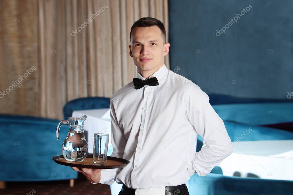 A young, handsome waiter is holding a tray in a carafe of water. The concept of the restaurant business.