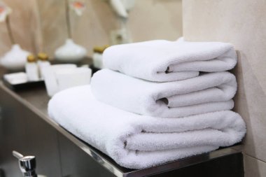 Three white bath towels are stacked on a bathroom shelf. A vase of flowers is out of focus. Hotel business concept. clipart