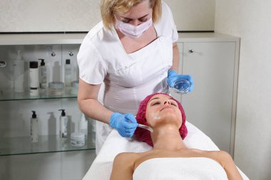 Bir kozmetikçi hastanın boynuna cildini beslemek, nemlendirmek ve tazelemek için bir ürün koyar. Klinikte estetik ameliyat. Hastanın yüzüne yakın çekim. Güzellik bürosu
