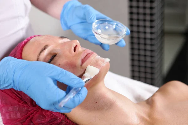 Primer plano de la cara del paciente en el consultorio de cosmetología. Aplicar en la cara significa hidratar y rejuvenecer la piel. lifting facial no quirúrgico. Cepillo con la droga en las manos de un esteticista . — Foto de Stock