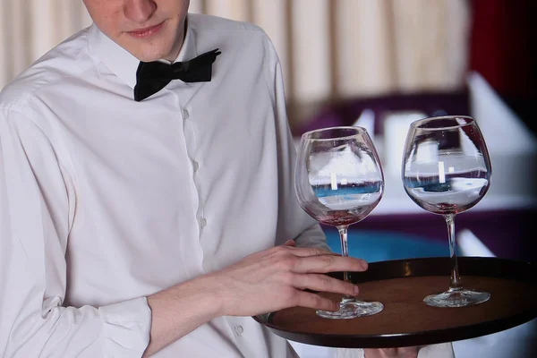 Un joven camarero un hombre con una camisa blanca trajo dos copas de vino para el vino en una bandeja. Foto sin rostro en el interior del restaurante . — Foto de Stock