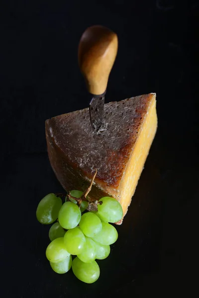 A large slice of hard cheese with a knife for cutting cheese and grapes on a black background. View from above. Isolated object. Craft cheese concept. Still life concept. — Stock Photo, Image