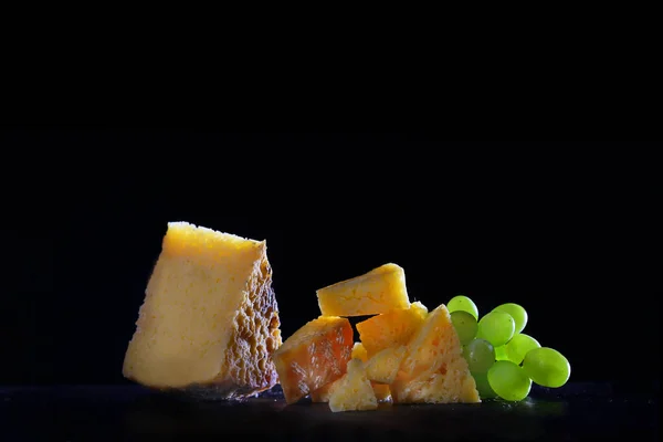 Pieces of cheese on a black background with grapes. Copy space. Still life on a black background. Homemade cheese concept. — Stock Photo, Image