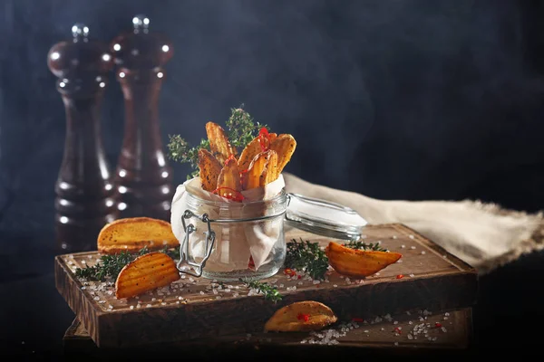 stock image Chips of fried potatoes in a glass jar on a black background. French fries on a wooden board with smoke. Still life concept. Copy of space.