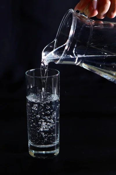 Female hand pours water from a glass decanter into a glass. Photo on a black background.