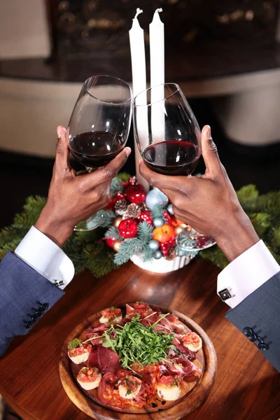 Two African American hands with glasses of red wine in focus. Hands raised on the background of the Christmas composition with candles. The concept of business meetings. New year.