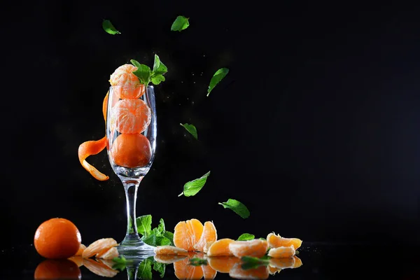 Tangerines with mint in a tall wine glass on a black background. Mint leaves fall on the steel, falling leaves. — Stock Photo, Image