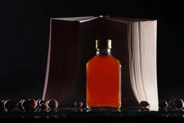A small bottle with a strong alcoholic drink of brown color stands on the background of an open book. The book is out of focus. Macro photo on black background with reflection. Hazelnuts are out of fo — Stock Photo, Image
