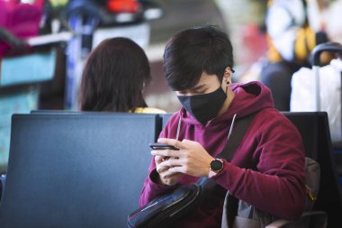 Yüzünde siyah bir maske olan Çinli bir adam havaalanında oturuyor ve elinde bir telefon tutuyor. Çin 'de Coronavirus. Salgın tehdidi.