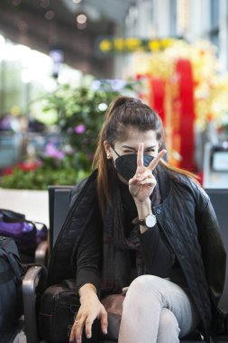 Yüzünde siyah tıbbi maske olan bir kız havaalanında oturuyor ve iki parmaklı bir işaret gösteriyor. Çin 'de Coronavirus. Salgın tehdidi.