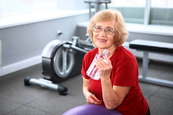 An elderly woman goes in for sports and holds a water bottle in her hands. Active and healthy lifestyle in old age. Photo in the interior. Copy space.