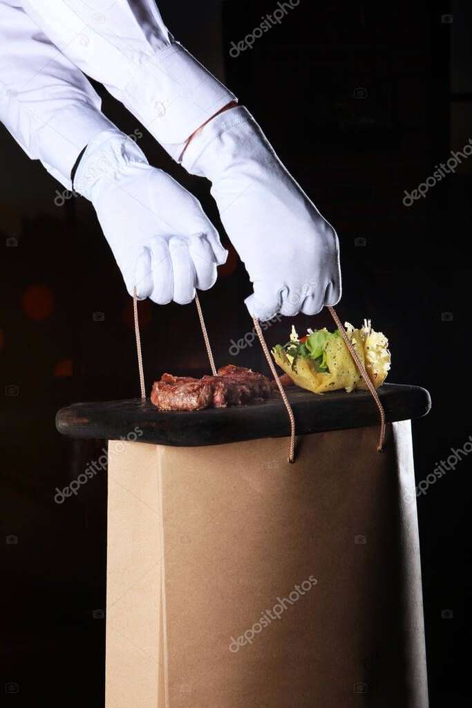 Food delivery to your home from the restaurant. White-gloved hands hold a bag of food. Dark background. Copy space.
