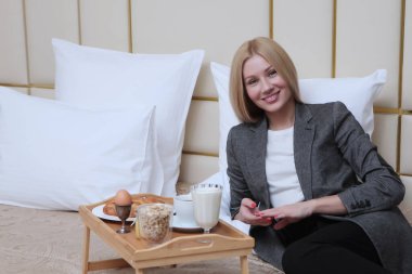 Ceketli genç bir kadın otel odasındaki bir yatakta kahvaltı yapıyor. Seyahat ve iş seyahatleri kavramı. Fotoğraf otelin içinde..