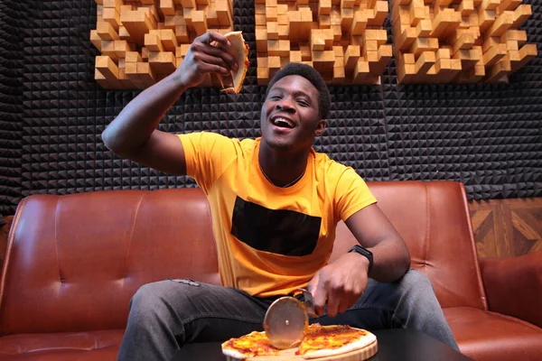 The cheerful African-American held up a slice of pizza. Photo in the interior. The concept of a good mood. Copy of the space.