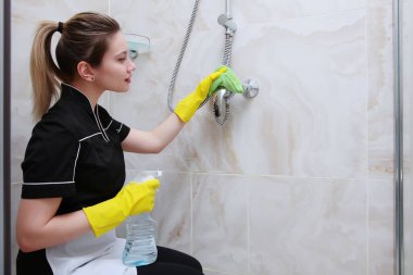 Üniformalı kız duş kabinindeki musluğu temizliyor. Peçete ve sprey hazır. Banyo mermer kiremitlerle süslenmiş..