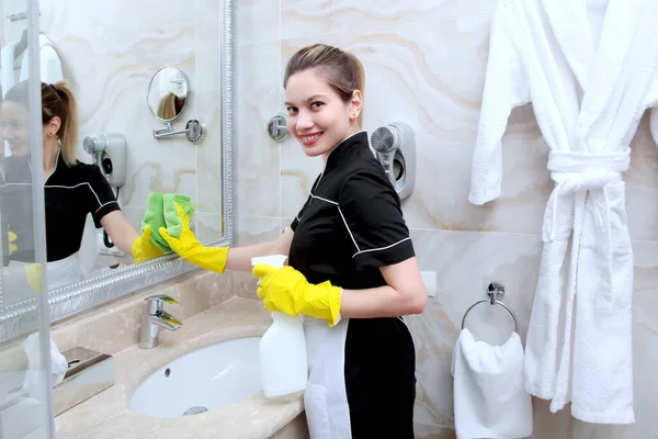 Üniformalı bir kız otel odasındaki aynayı temizliyor ve gülümsüyor. Otel işi kavramı.. — Stok fotoğraf
