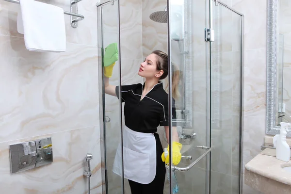 Una joven en uniforme lava las puertas transparentes en la cabina de ducha. Servilleta y spray en la mano. El concepto de limpieza e higiene . — Foto de Stock